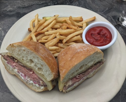 A plate with a sandwich filled with meat and cheese, served with fries and a small bowl of ketchup.
