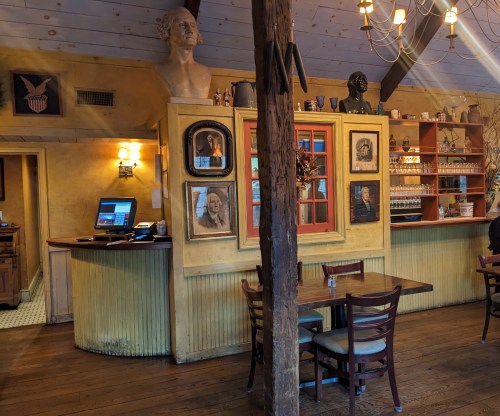 Cozy restaurant interior with wooden tables, vintage decor, and busts on display against a warm yellow wall.
