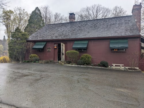 A red wooden house with green awnings, surrounded by trees, on a rainy day. The driveway is wet and reflective.