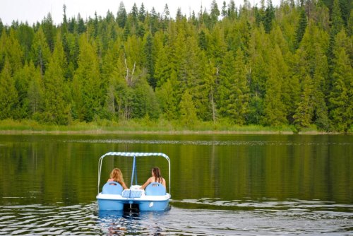 Two people paddle a blue boat on a calm lake surrounded by lush green trees.