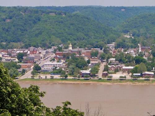 A scenic view of a small town along a river, surrounded by green hills and trees on a sunny day.