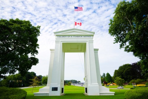 White archway with flags of the USA and Canada, inscribed "Children of a Common Mother," surrounded by greenery.