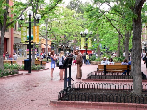 A bustling outdoor plaza with trees, benches, and people socializing on a sunny day.