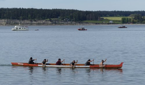 A group of six people paddling a canoe on calm water, with boats and trees in the background.