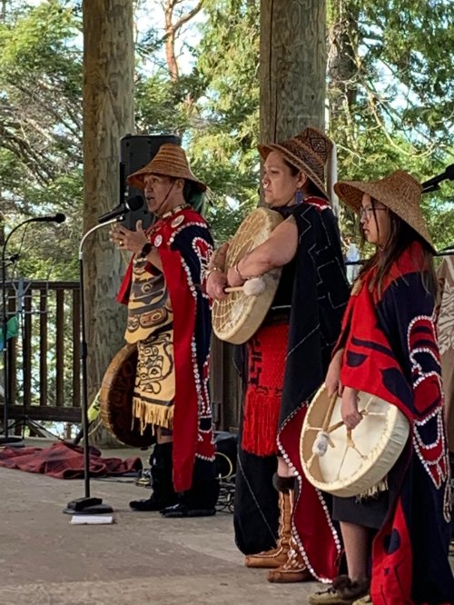 Indigenous performers in traditional attire, holding drums, speaking at a cultural event outdoors.