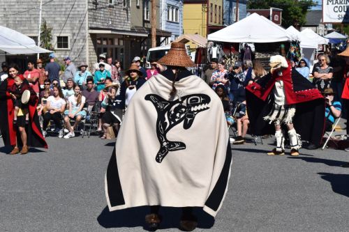 A performer in a traditional cape with a whale design stands before an audience at a cultural festival.