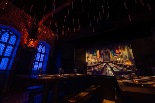 A dimly lit dining hall with long tables, candle-like lights hanging, and a large screen depicting a grand hall.
