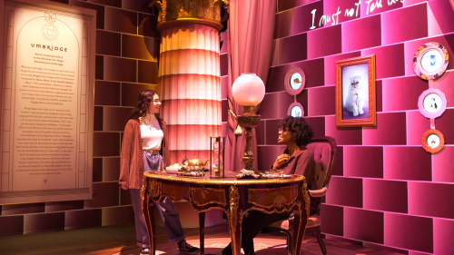 Two women interact in a whimsical room with ornate decor, a table, and framed art on the walls.