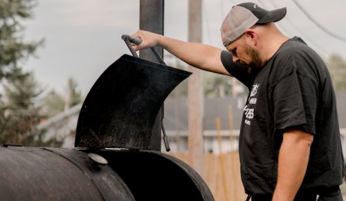 A man in a cap checks a smoker, releasing smoke as he lifts the lid.