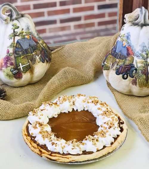 A pumpkin pie topped with whipped cream and nuts, surrounded by decorative pumpkins and burlap.