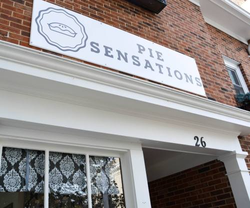 Sign for "Pie Sensations" on a brick building, featuring a pie graphic and decorative window lace.