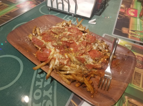 A wooden platter of loaded fries topped with melted cheese, pepperoni, and herbs, served with a fork.