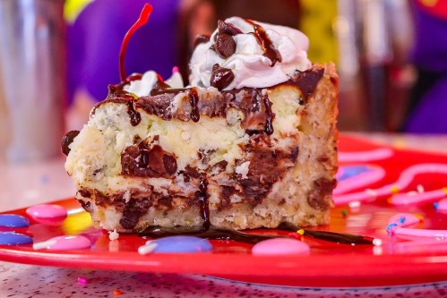 A slice of chocolate chip cheesecake topped with whipped cream, chocolate drizzle, and a cherry on a colorful plate.