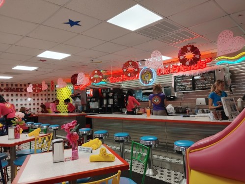 A colorful diner interior with retro decor, bright neon signs, and staff serving customers at the counter.