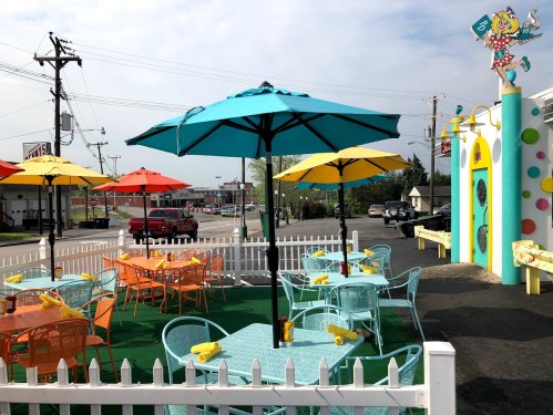 Colorful outdoor dining area with blue and yellow umbrellas, orange and teal tables, and a white picket fence.