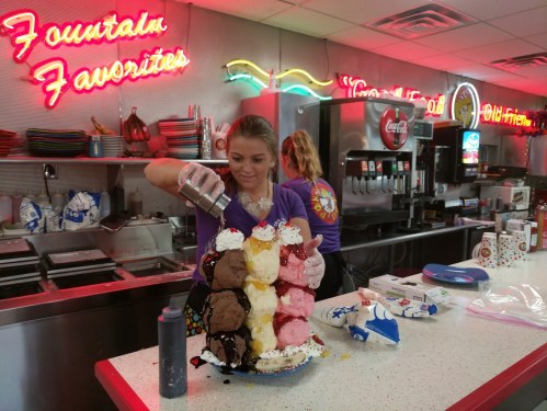 A woman prepares a towering ice cream sundae with various flavors and toppings at a retro diner. Neon signs glow in the background.
