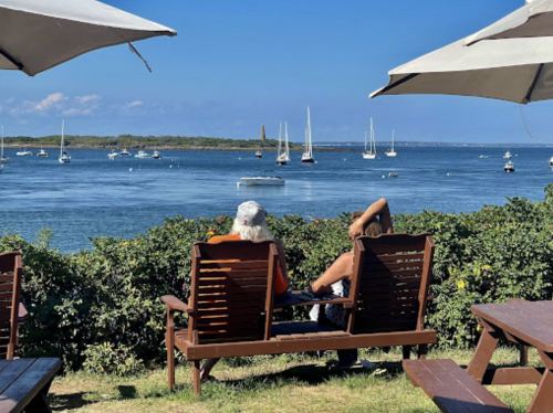 Two people relax on wooden chairs by the water, with sailboats and a clear blue sky in the background.
