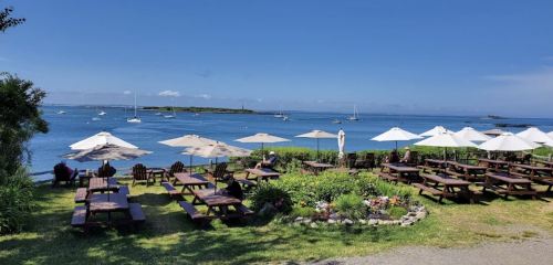 A scenic view of a waterfront restaurant with picnic tables, umbrellas, and boats in the calm blue water.