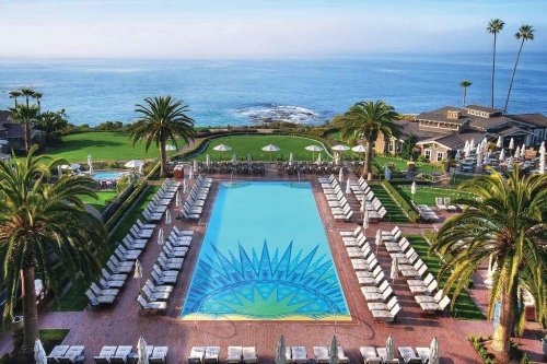 A scenic view of a pool surrounded by palm trees, overlooking the ocean with lounge chairs and a lush green lawn.