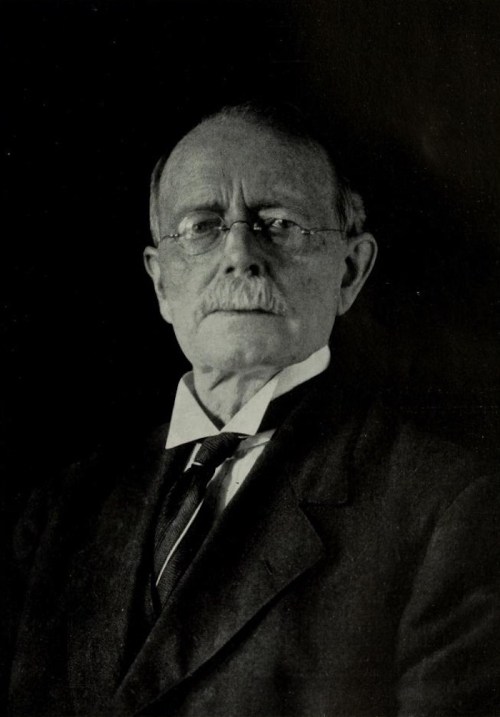 A portrait of an older man with glasses, a mustache, and a formal suit, set against a dark background.