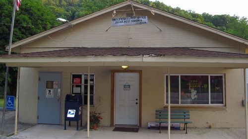 A small, beige post office building with a sloped roof, featuring a blue door and a bench outside.