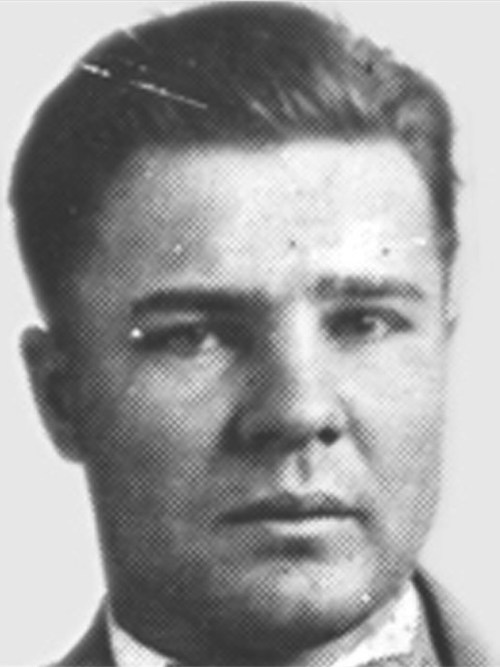 Black and white portrait of a man with short hair, wearing a suit and tie, looking directly at the camera.
