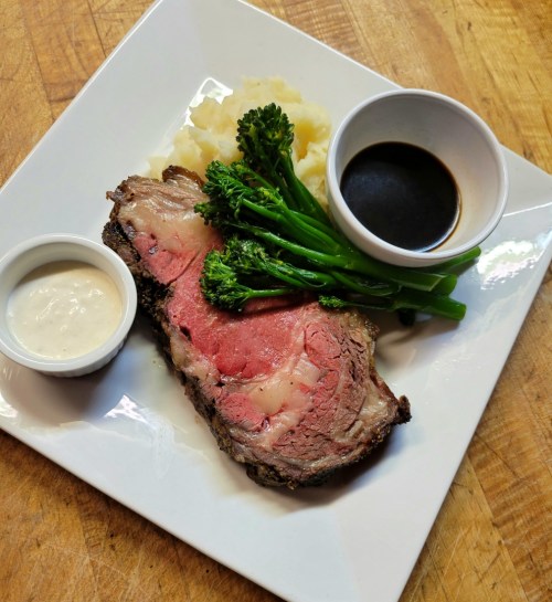 A plate featuring sliced roast beef, mashed potatoes, broccolini, and two dipping sauces.