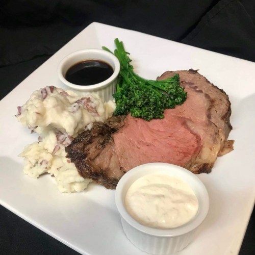 A plate featuring a slice of prime rib, mashed potatoes, broccolini, and two dipping sauces.