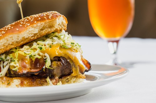 A juicy burger with cheese, lettuce, and bacon on a sesame seed bun, served with a glass of amber beverage in the background.