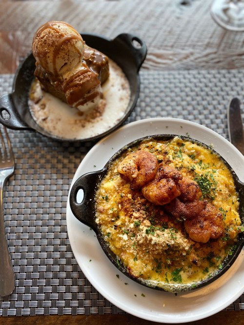 A cast iron dish with shrimp and cheesy casserole, alongside a dessert of ice cream and caramel drizzled on a cake.