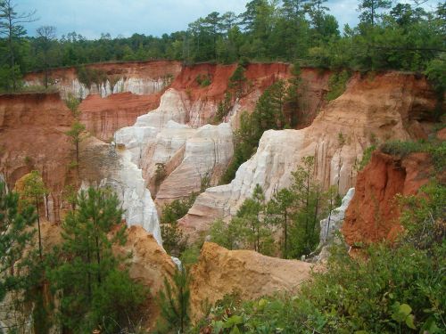 A colorful canyon with red and white cliffs, surrounded by green trees under a cloudy sky.