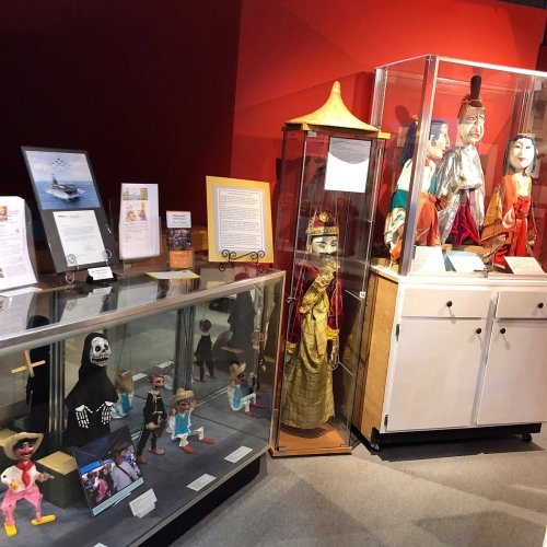 Display of colorful puppets and artifacts in a museum setting, with a red wall backdrop and glass cases.