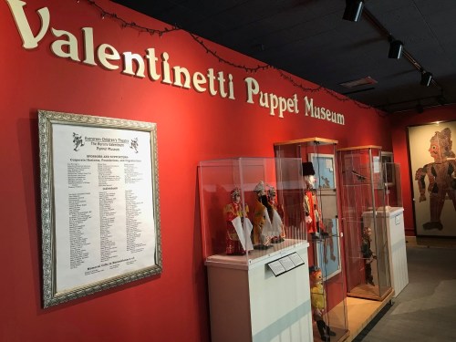 Interior of the Valentinetti Puppet Museum, featuring display cases with puppets and a list of performers on the wall.