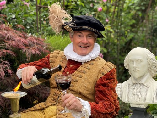 A man in historical costume pours wine while seated in a garden, with a bust of Shakespeare nearby.