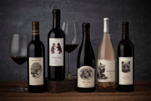 A selection of six wine bottles with artistic labels, accompanied by two wine glasses on a wooden table.