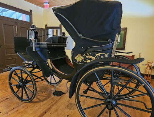 A vintage black horse-drawn carriage with ornate details, displayed indoors on a wooden floor.