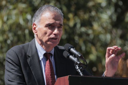 A man in a suit speaks at a podium with microphones, gesturing with his hand against a blurred outdoor background.