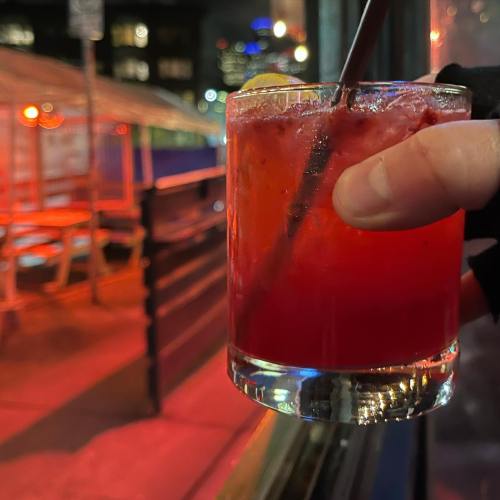 A hand holding a vibrant red cocktail with a straw, set against a nighttime urban backdrop with glowing lights.