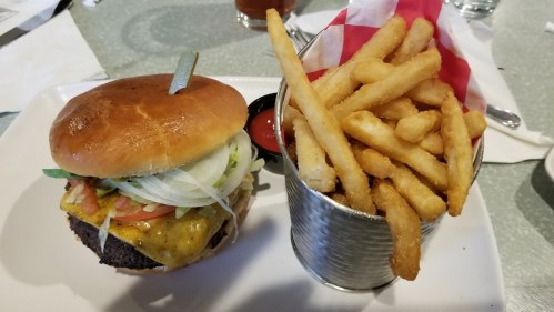 A juicy cheeseburger with lettuce, tomato, and onion, served with a side of crispy French fries in a metal cup.
