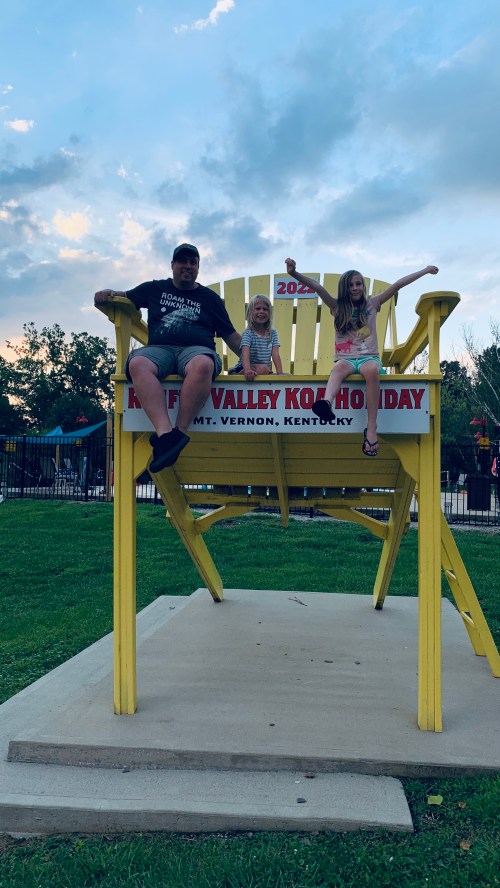 A man and two girls sit on a large yellow chair at a fair, smiling and enjoying the outdoor setting.