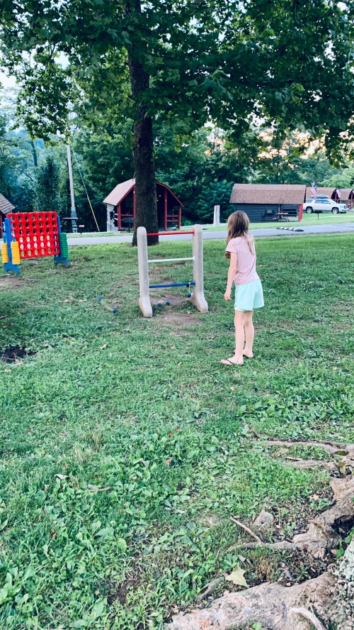 A child stands in a grassy area near playground equipment, looking towards a colorful game structure in the background.
