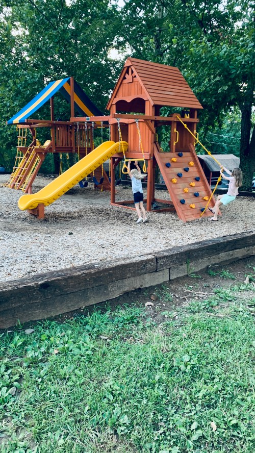 Two children play on a colorful wooden playground with a slide, climbing wall, and swings, surrounded by trees.