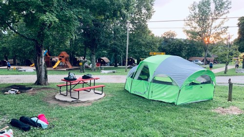 A green and gray tent set up in a grassy campsite near a playground at sunset, with a picnic table nearby.