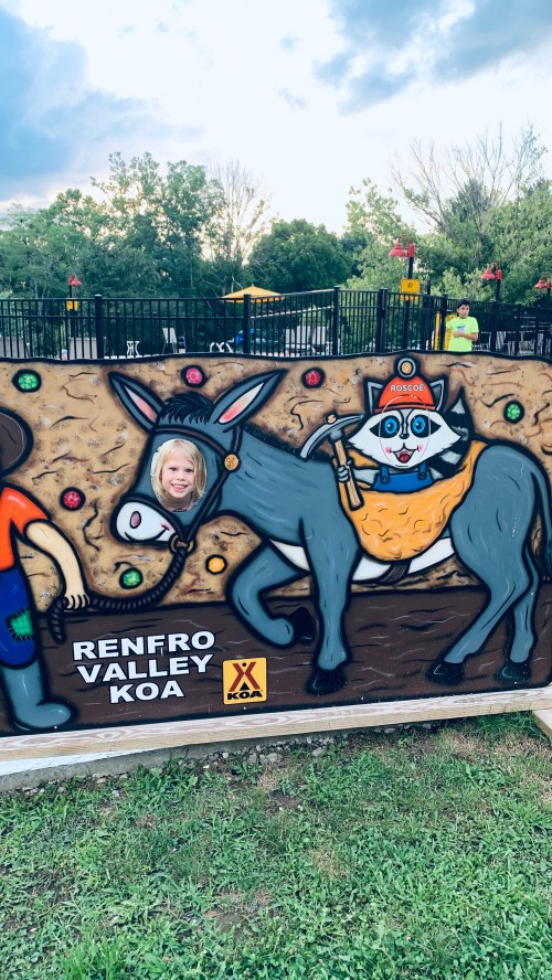A child peeks through a cutout of a donkey in a colorful mural at Renfro Valley KOA, with playful characters around.