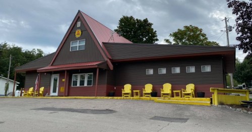 A brown and red building with a triangular roof, featuring yellow chairs on the porch and an American flag.