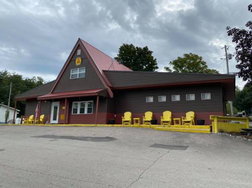 A brown and red building with a triangular roof, featuring yellow chairs on the porch and a cloudy sky above.
