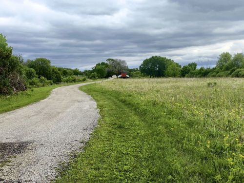 A winding gravel path leads through a grassy field under a cloudy sky, with trees and a small red building in the distance.