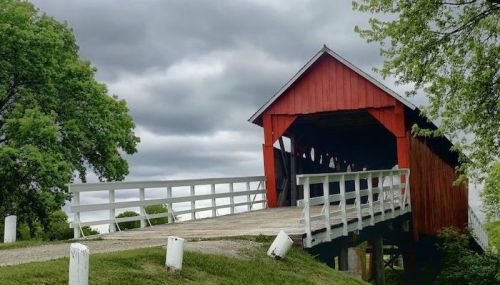 A red covered bridge with a white railing, set against a cloudy sky and surrounded by green trees.