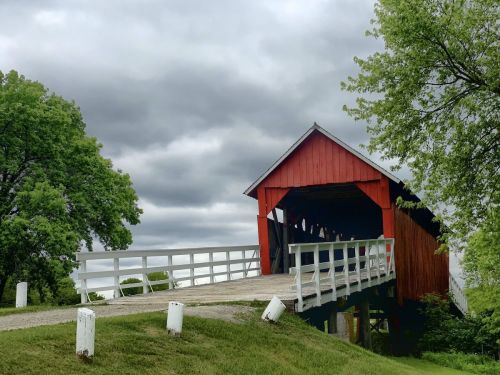 A red covered bridge stands over a grassy area, surrounded by trees under a cloudy sky.