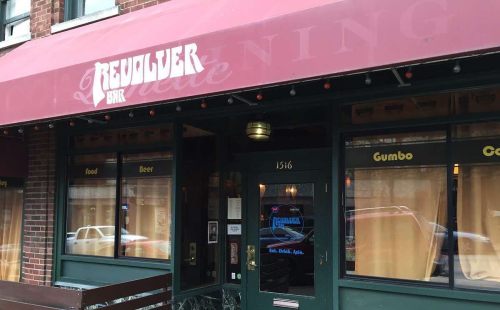 Exterior of the Revolver Bar, featuring a red awning and windows with menu items like gumbo and beer.